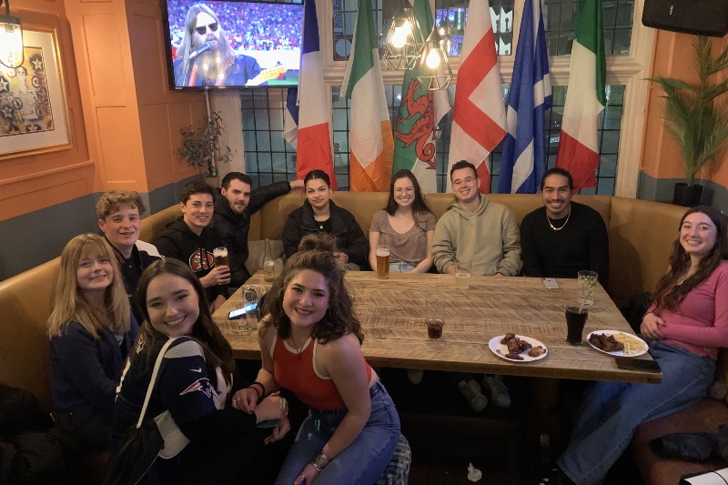 University of Bristol students gather around a table in a pub with the Super Bowl on a television in the background.
