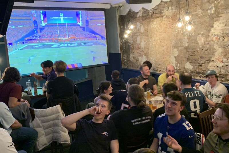 University of Bristol students gather in a pub with the Super Bowl on a projector in the background.
