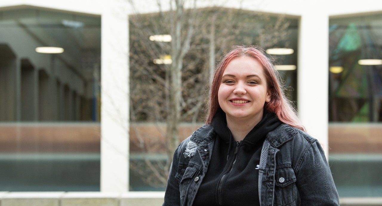Futures Scholar Maja is smiling at the camera. She is wearing a black hooded sweatshirt and dark denim jacket, and is standing in front of a tree and a building with white pillars.