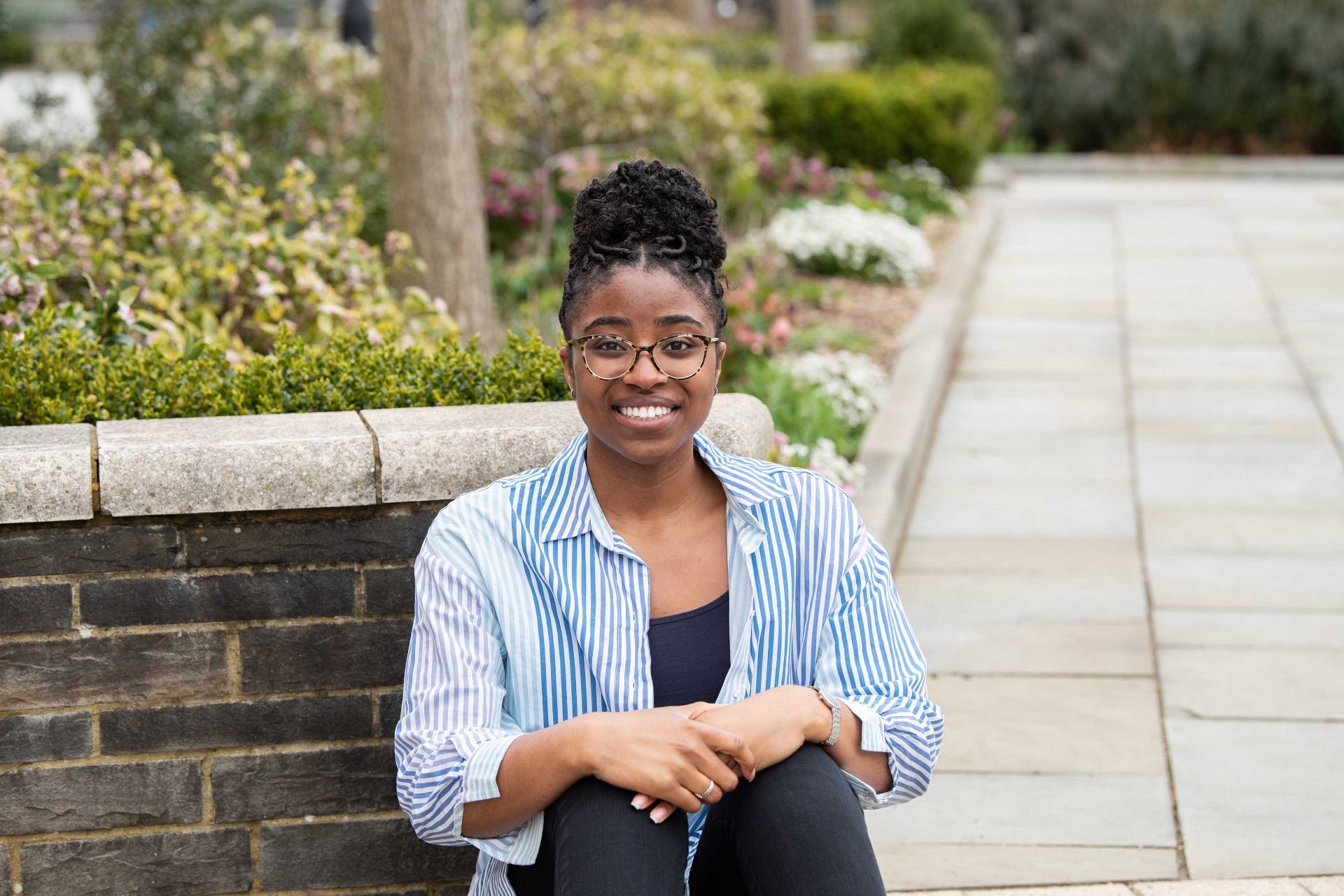 University of Bristol student Chinelo Etiaba wears professional attire as she smiles, sitting in a garden with a stone wall behind her.