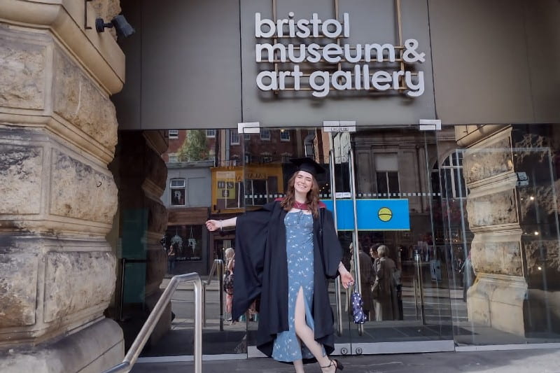 Helen celebrates her graduation with a smile, as she stands outside the Bristol Museum and Art Gallery, dressed in her cap and gown.