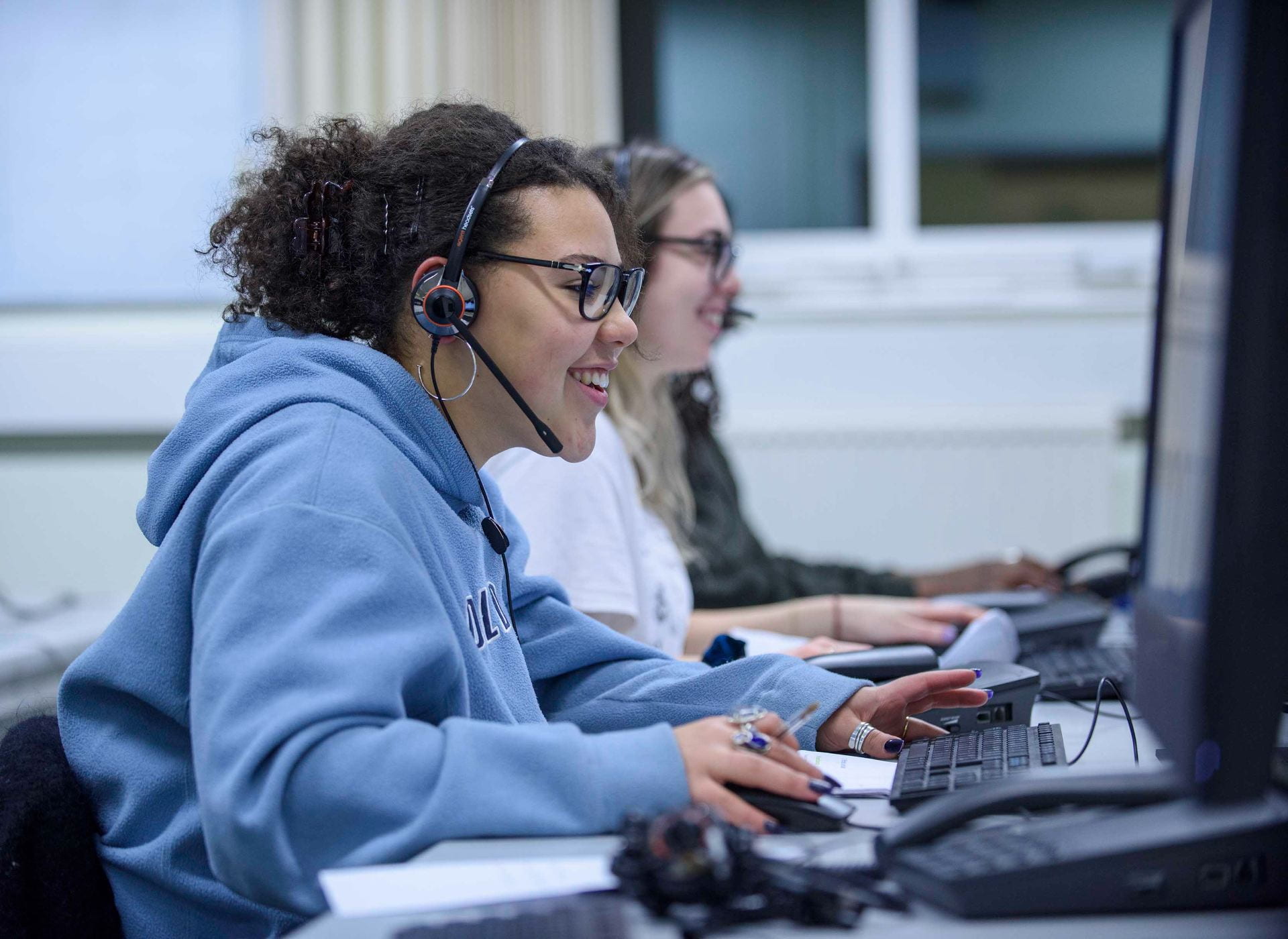 Two student callers wearing headsets smile and use a computer.