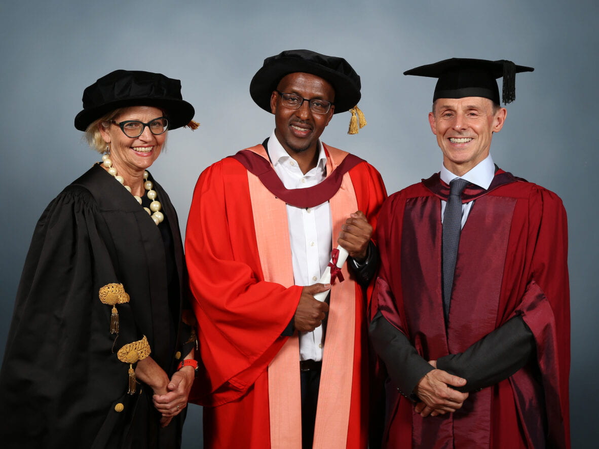 Latif Ismail poses in honorary graduate gown while holding degree. Professor Professor Judith Squires stands on the left and Professor Eric Herring stands on the right, both wearing ceremonial robes.