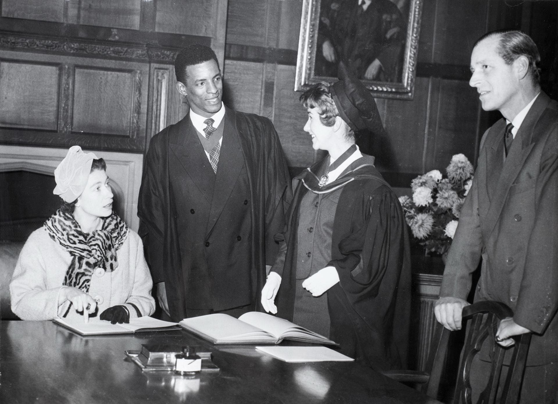 George Odlum stood in conversation with a fellow student (both wearing formal gowns) and Prince Andrew, Queen Elizabeth II is seated at the table beside Odlum.