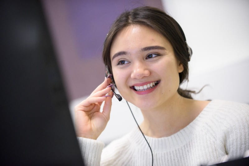 A student is smiling as she adjusts her computer headset while on a telethon call to an alum. She is wearing a white top, and is looking away in the distance to her right.